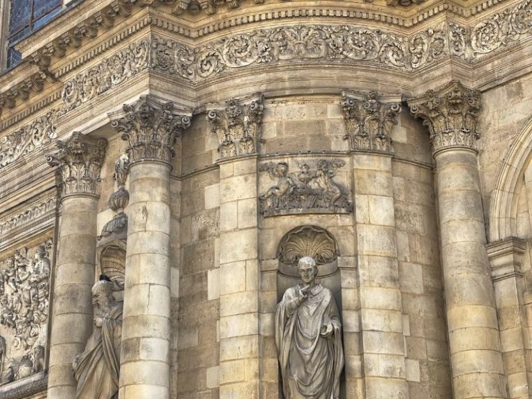 Stone statues of robed figures and cherubs adorn the ornate facade of a classical building with Corinthian columns.