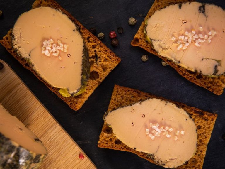 Three slices of foie gras on toast, sprinkled with sea salt and peppercorns, on a dark surface.