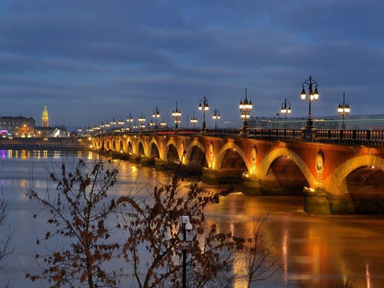 A wide brick bridge with arched spans is illuminated by ornate lampposts and reflects in the water at dusk.