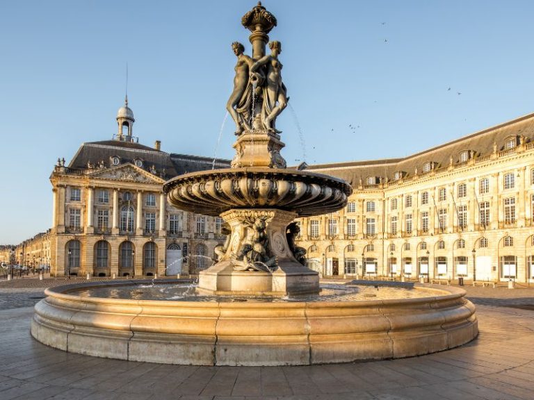 Grand fountain with three nude figures at the top, spraying water, in front of ornate classical buildings.