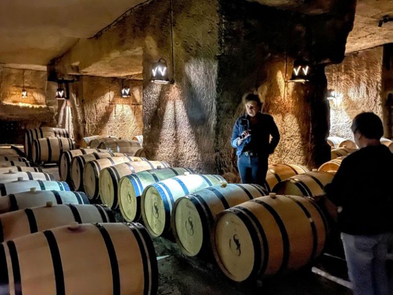A woman in a denim jacket stands in a cellar, gesturing towards rows of wine barrels. Another person is visible from behind, facing the barrels.