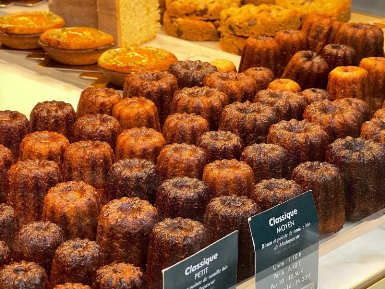 Rows of golden-brown canelés, a French pastry, with some larger cakes in the background. Labels indicate 'Classique MOYEN' and 'Classique PETIT'.