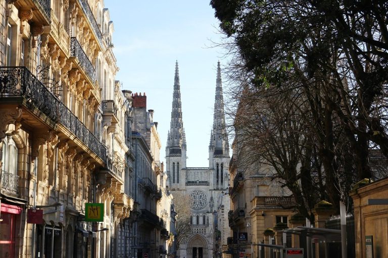 A street view leading to a large cathedral with twin spires, flanked by ornate buildings and bare trees.
