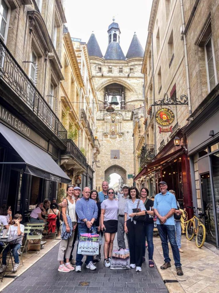 A group of eleven people pose for a photo on a cobblestone street in front of the Grosse Cloche bell tower in Bordeaux, France.