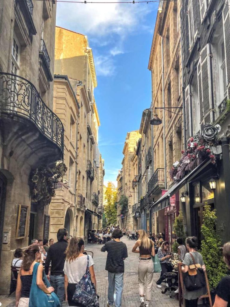 People stroll down a narrow European street lined with tall buildings housing cafes and shops. Outdoor seating is occupied.