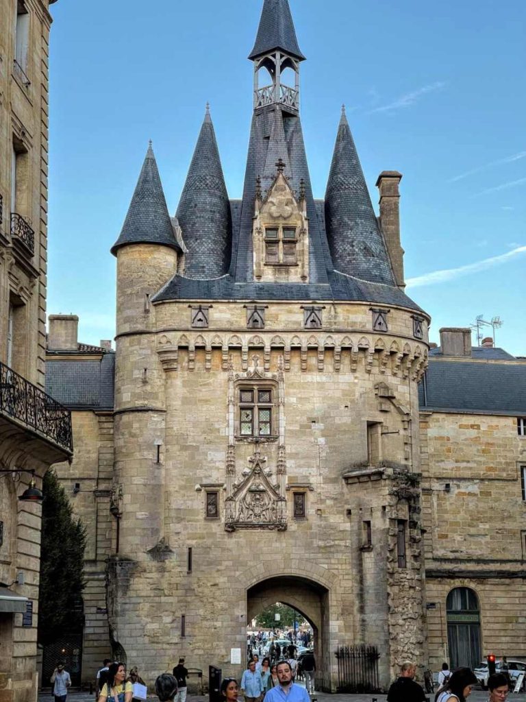 Gothic stone gate tower with conical roofs and a central arched passage. People walk through the archway.