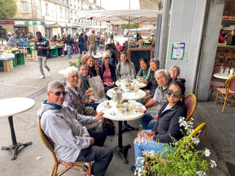 A group of nine people sit at tables outside a cafe, with a bustling market in the background.