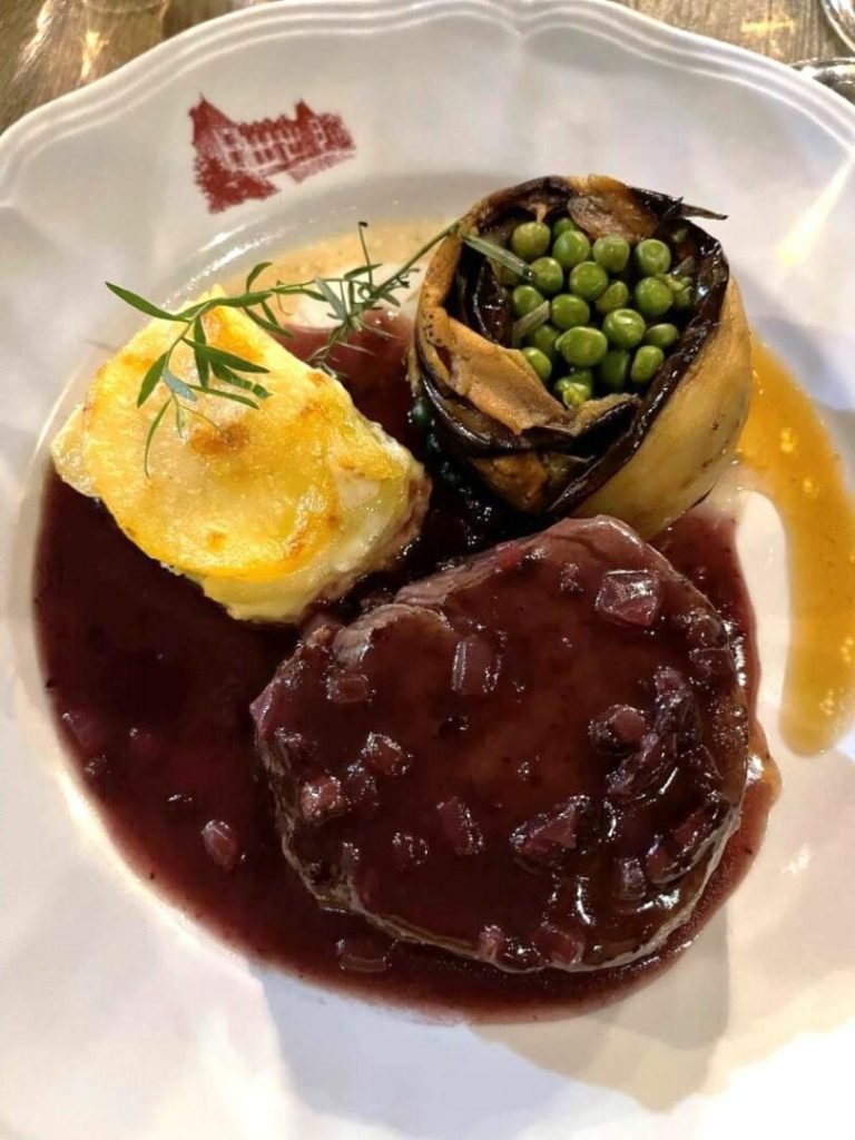 A plate of food featuring a steak in red wine sauce with onions, a slice of gratin potato, and a vegetable cup filled with peas.