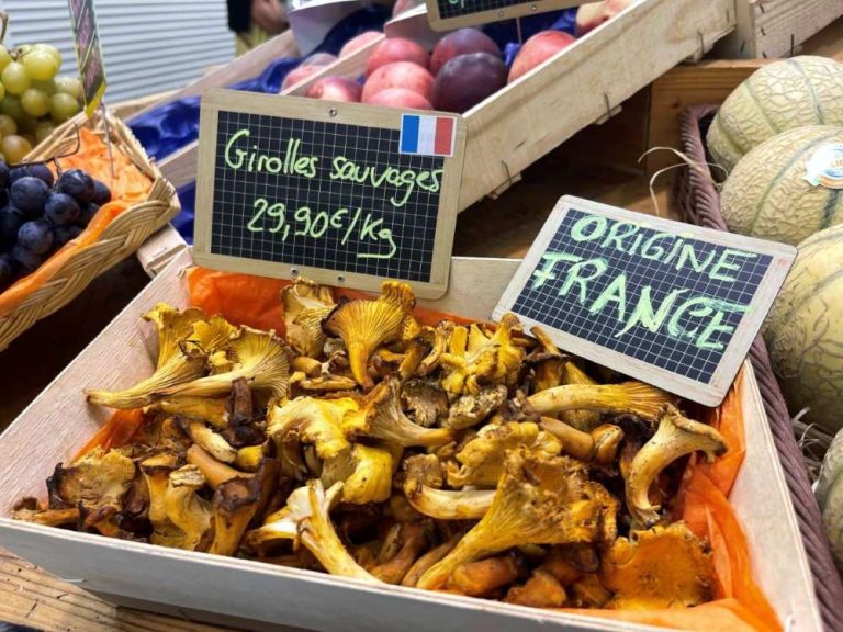 A wooden crate filled with fresh chanterelle mushrooms, with a chalkboard sign reading 'Girolles sauvages 29,90€/Kg' and another reading 'Origine France'.