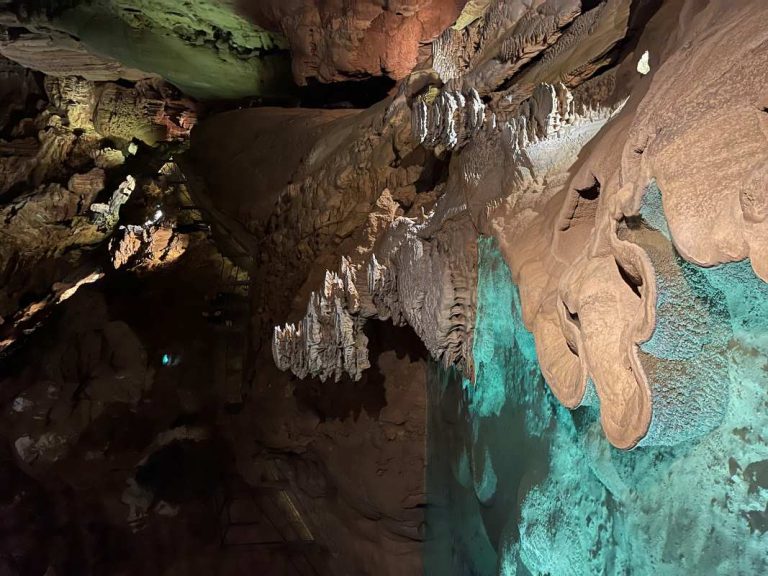 Cave formations with stalactites hanging from the ceiling and a turquoise pool of water reflecting the formations.