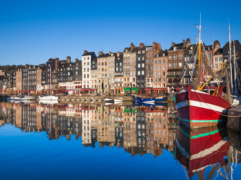 Colorful buildings line a harbor with boats, a red and white fishing boat in the foreground, all reflected in the blue water.