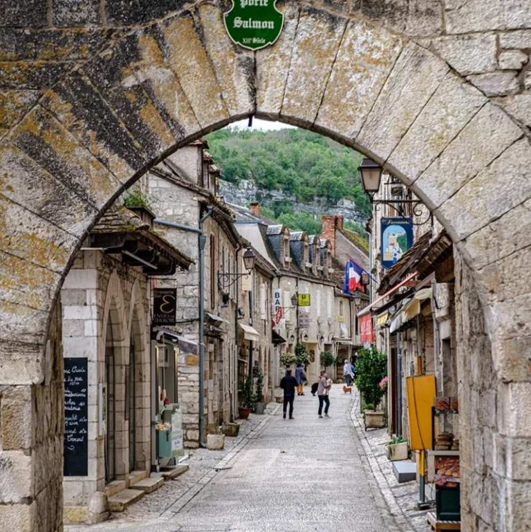 Stone archway framing a charming European street lined with shops and buildings. A sign reads 'Porte Salmon'.