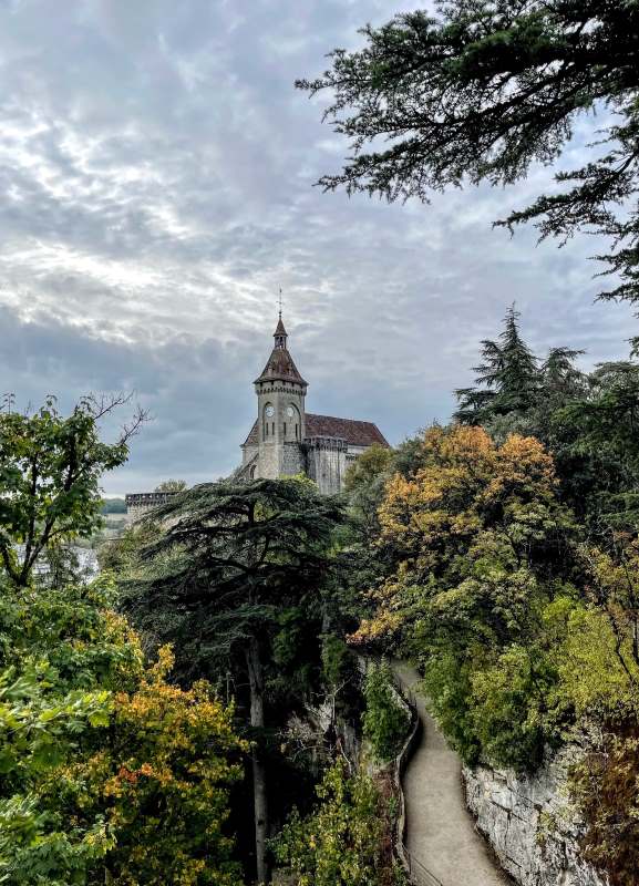 Stone church with a clock tower rises above lush green and autumn foliage, with a winding path leading through trees.