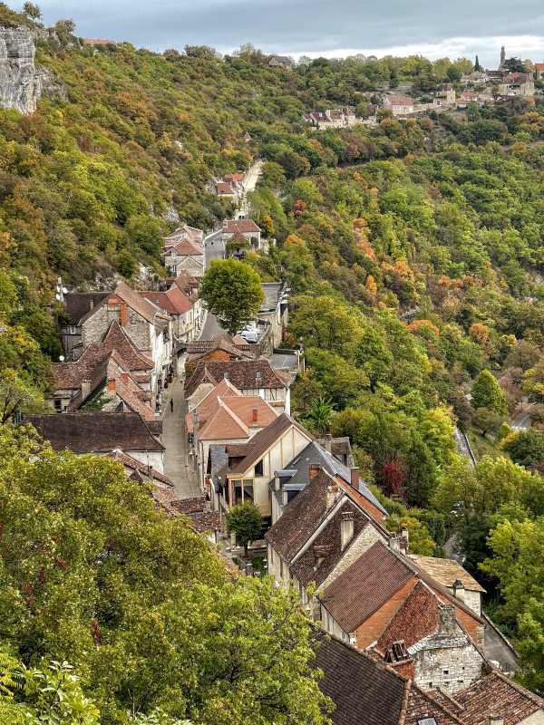 A village with stone buildings and terracotta roofs nestled on a steep, tree-covered hillside.
