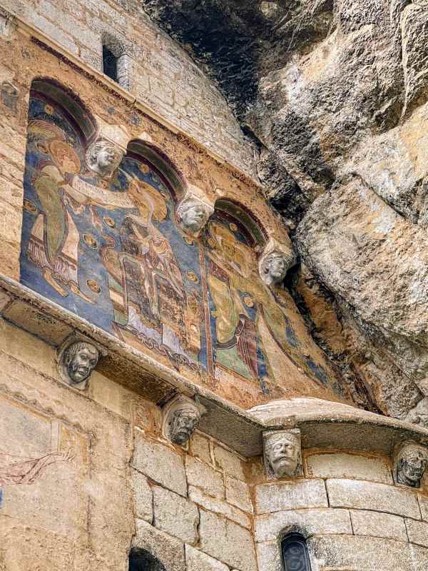 Ancient fresco depicting figures under arches, with carved faces adorning the stone building nestled into a rocky cliff.