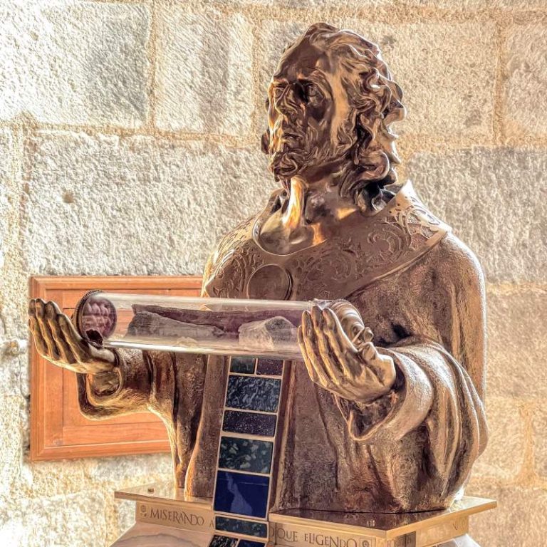 Bronze bust of a man with curly hair and beard, holding a glass reliquary with bones and a purple orb.