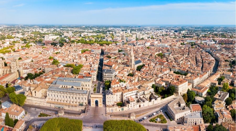 Aerial view of a historic European city with terracotta roofs, a prominent triumphal arch, and a tall church spire under a clear blue sky.