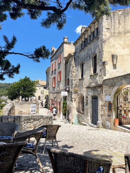 Cobblestone street in a European village with stone buildings, red shutters, and outdoor cafe seating in the foreground.