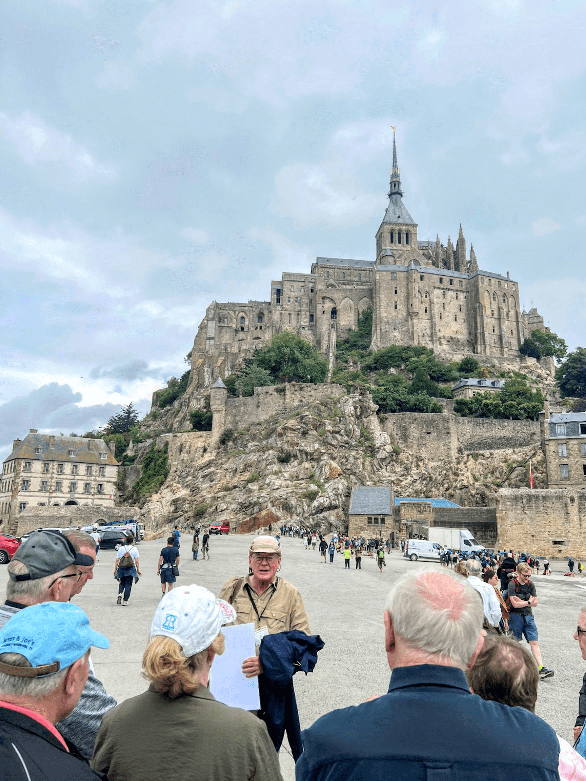 A tour guide addresses a group of tourists in front of the historic Mont Saint Michel abbey perched on a rocky island.
