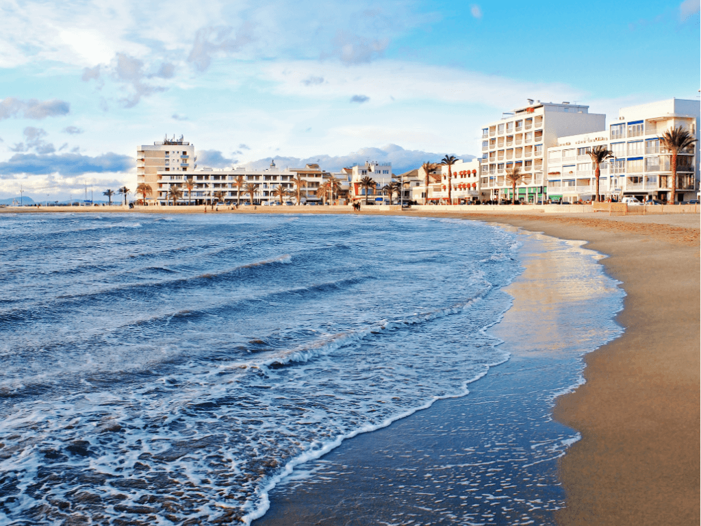 Gentle waves lap onto a sandy beach lined with palm trees and modern buildings under a partly cloudy sky.