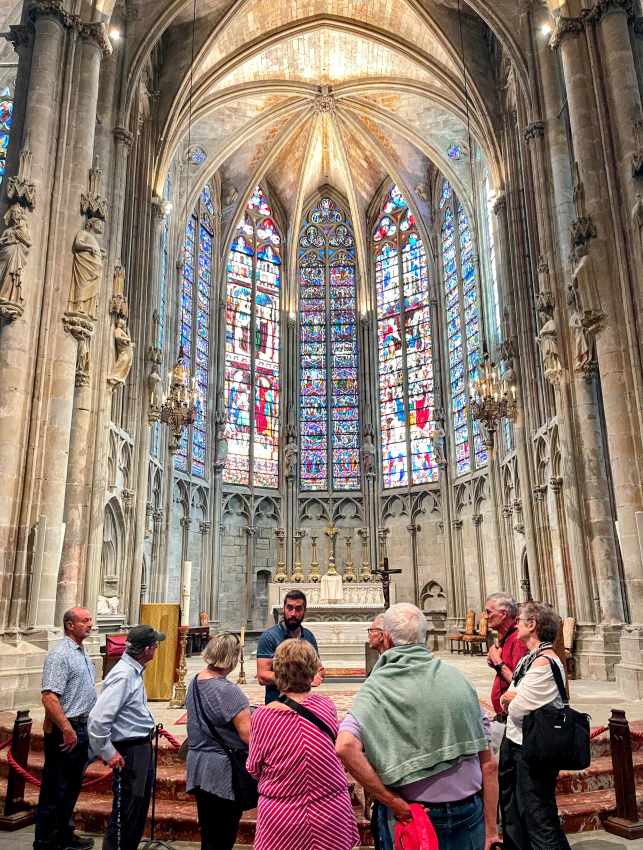 A tour guide speaks to a group of visitors inside a grand cathedral with ornate stained-glass windows and high vaulted ceilings.