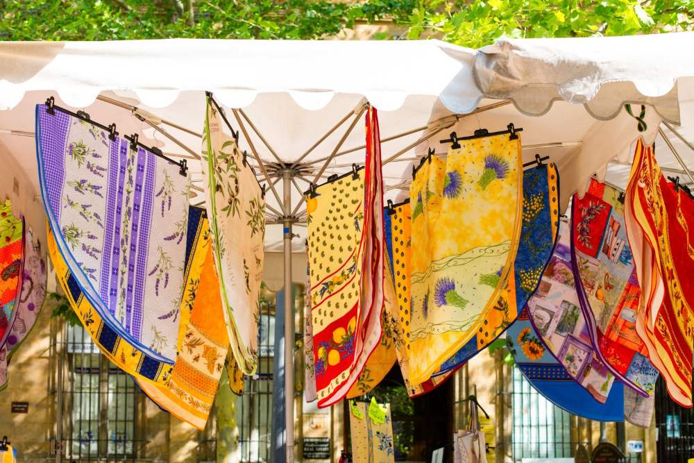 Colorful fabric squares with lavender, olive, fruit, and floral patterns hang from a market tent.