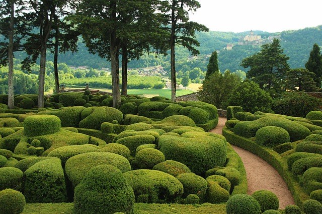 Topiary garden with sculpted green bushes and trees, overlooking a village and chateau in the distance.
