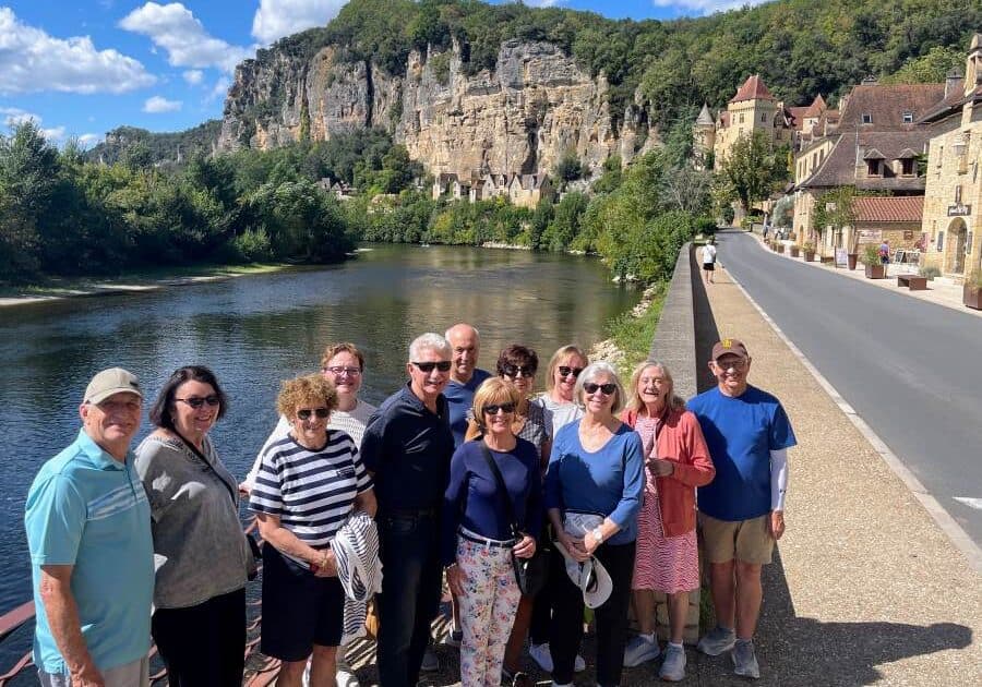 A group of ten adults stand on a stone walkway beside the Dordogne river with a scenic cliffside village in the background.