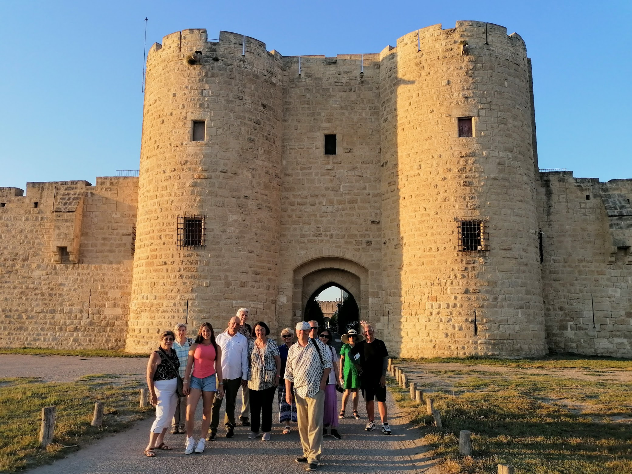 A group of people stand in front of a large stone medieval fortress gate on a sunny day.