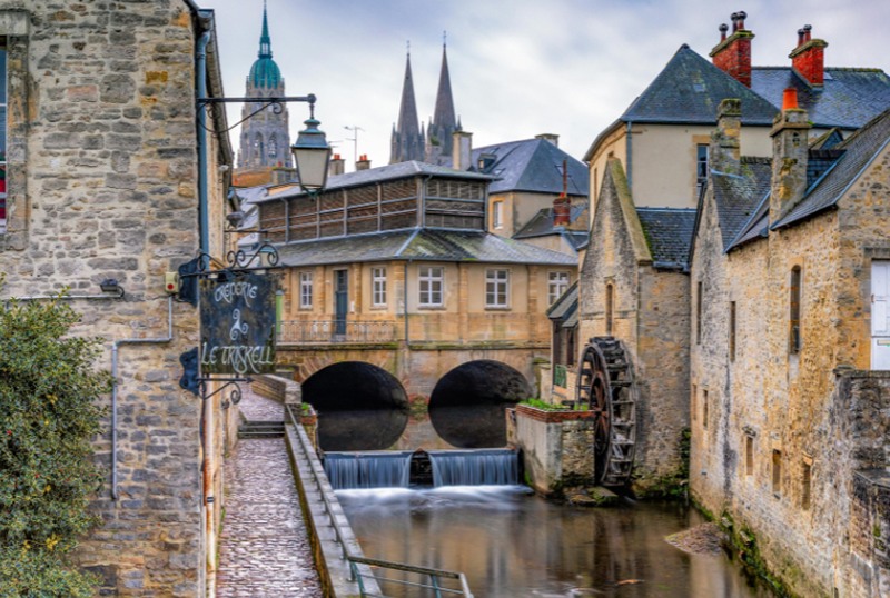 Creperie Le Triskell sign by a canal with a water wheel and bridges, medieval buildings, and distant church spires