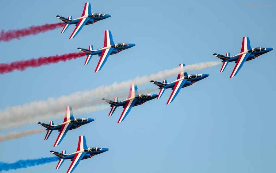 Several French Air Force jets flying in formation against a clear blue sky, trailing red, white, and blue smoke.