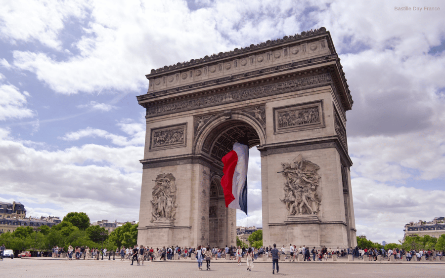 The Arc de Triomphe in Paris with a large French flag hanging from its arch, under a cloudy sky.