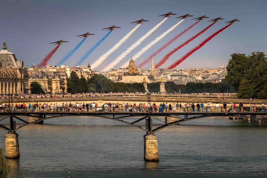 Jets flying over bridge in Paris with multicolored smoke trails, people look up from bridge