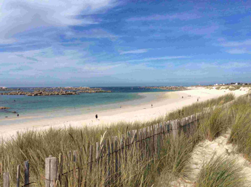 Sandy white beach, blue water with rock formations, tall grass and wooden fence in foreground