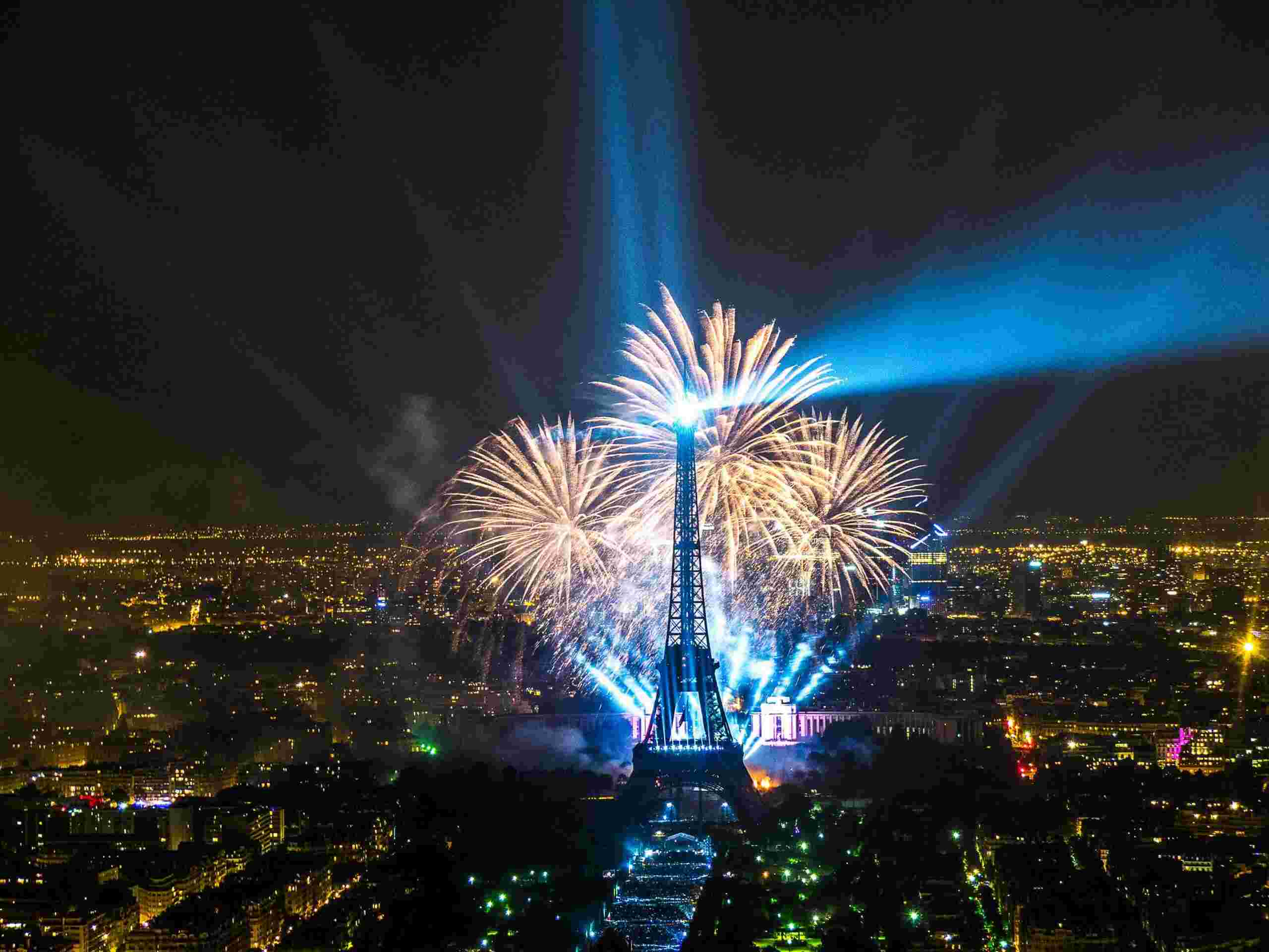 Aerial view of fireworks at night behind the Eiffel tower, sprawl of city lights behind