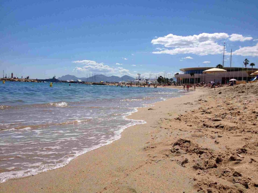 View of waterline and beach from sand level, glistening water, marina buildings and boats in background