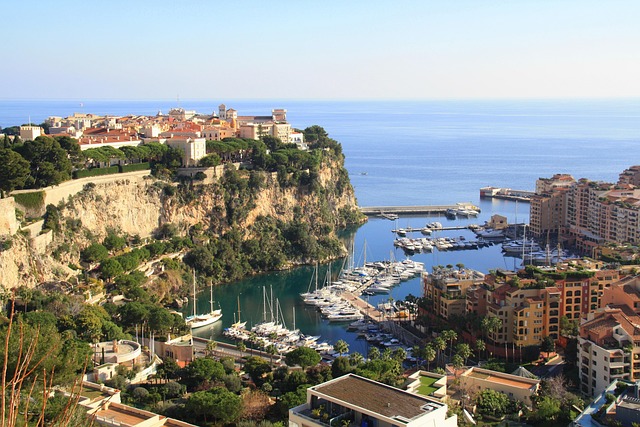 A wide view of a harbor filled with yachts, with a cliff-top town overlooking the water and the Mediterranean Sea.