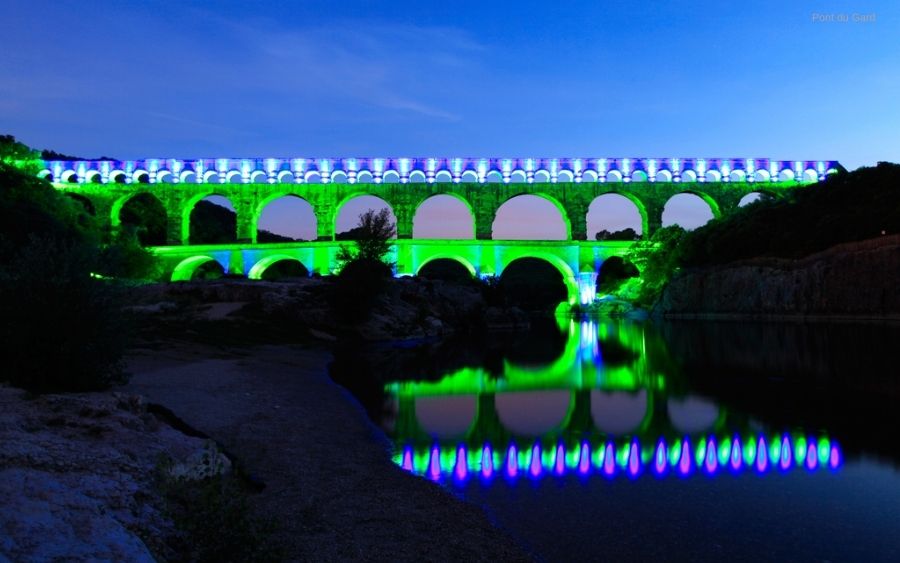 Pont du Gard aqueduct illuminated with green and blue lights at night, reflecting in the water.