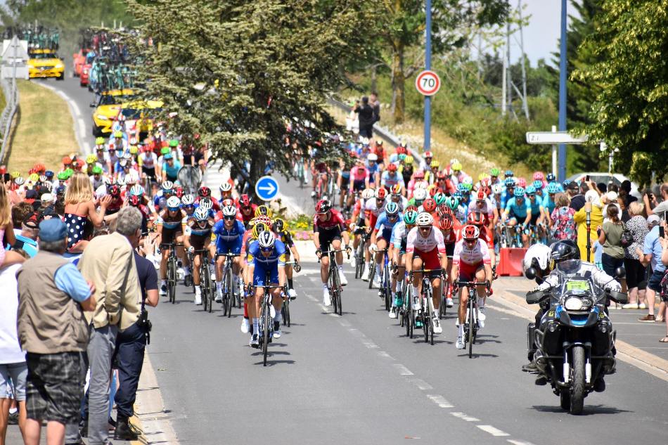 A large peloton of cyclists races down a road lined with spectators. A police motorcycle escorts the riders.