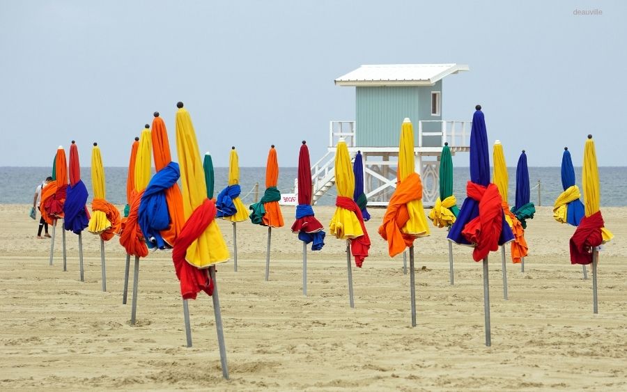Colorful closed beach umbrellas stand in rows on a sandy beach with a light blue lifeguard station in the background.