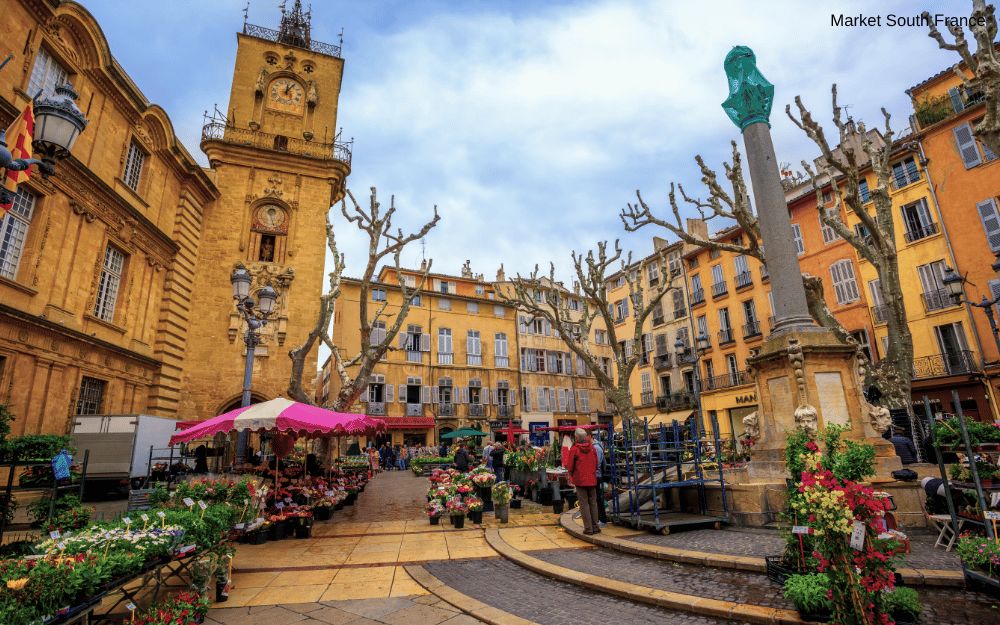 A bustling market square in a South of France town with a clock tower and flower stalls