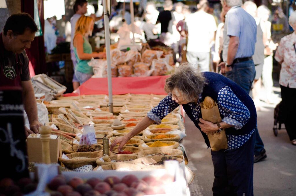 Old woman holding baguette leaning over to view a market vendor's product