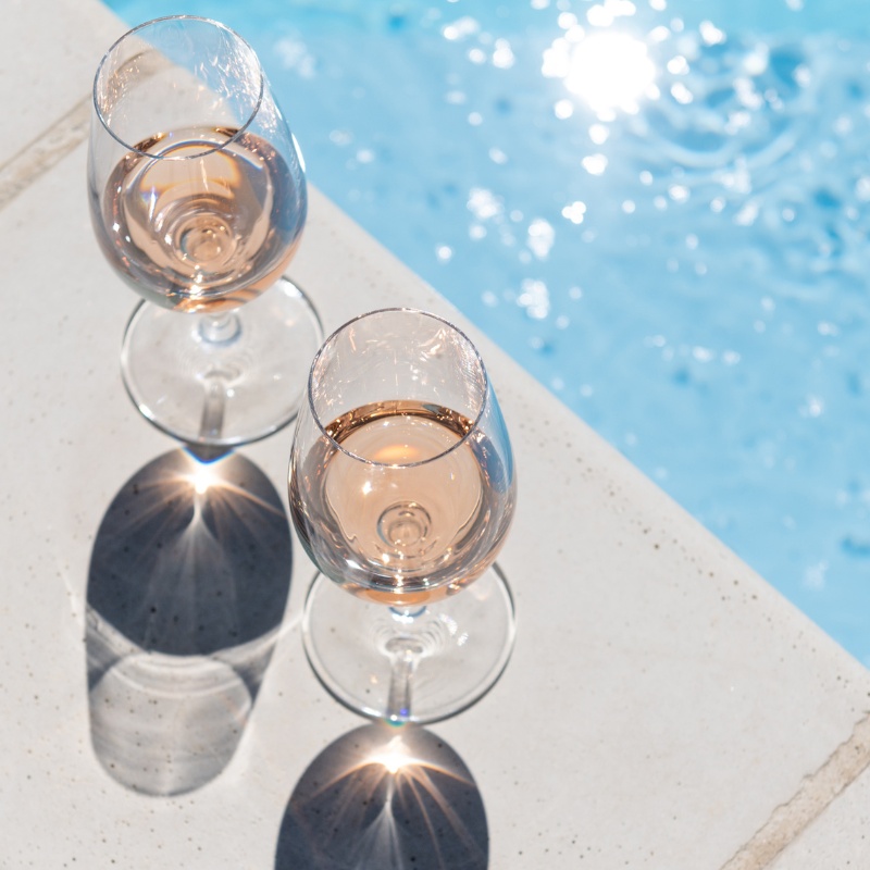 Two glasses of pale rosé wine on the stone edge of a swimming pool, sun reflecting in water and casting shadows of the glasses