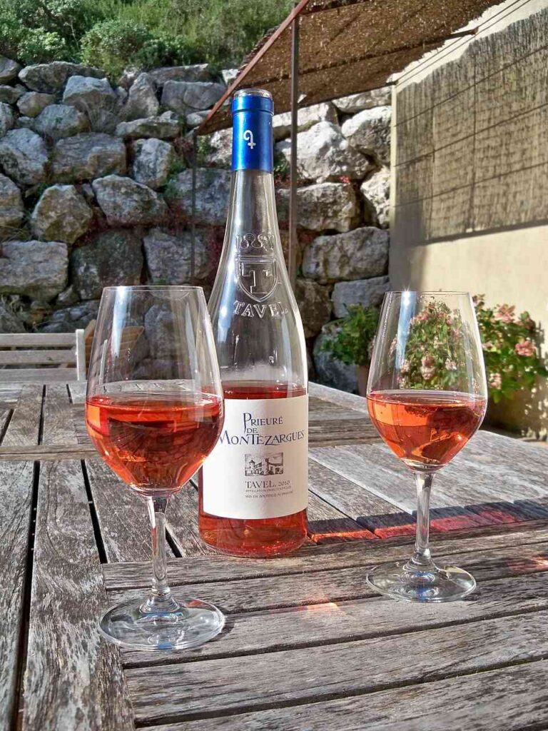A glass of deep pink Tavel on a weathered wooden table with stone wall and awning in background, evocative of a patio in Provence