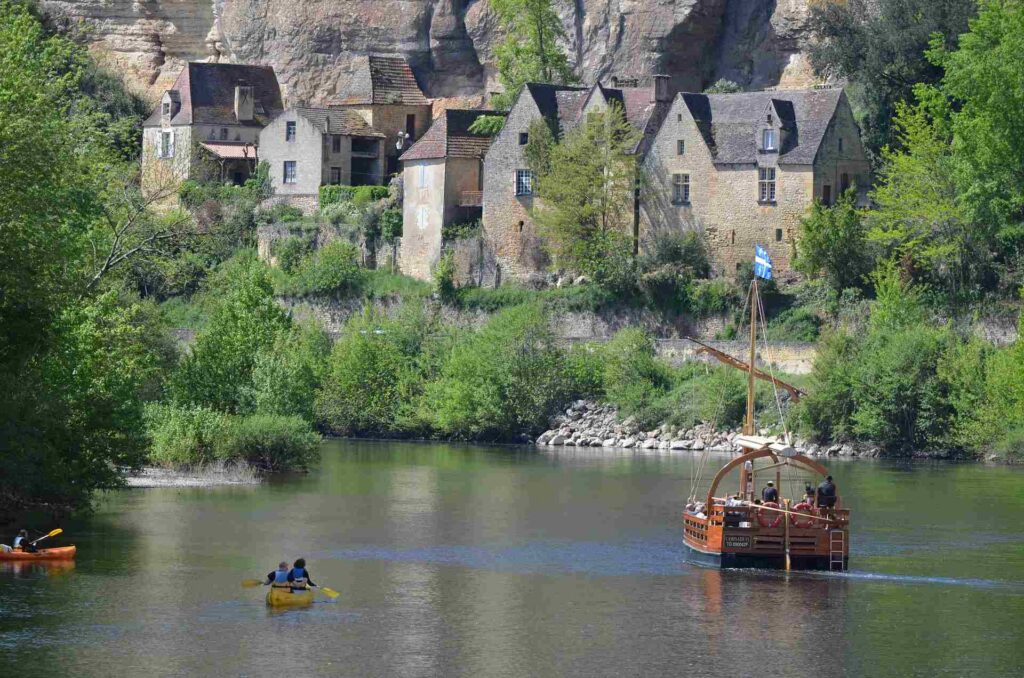 A traditional gabarre and canoes on the Dordogne River below the stone houses of La Roque-Gageac, set against the cliff